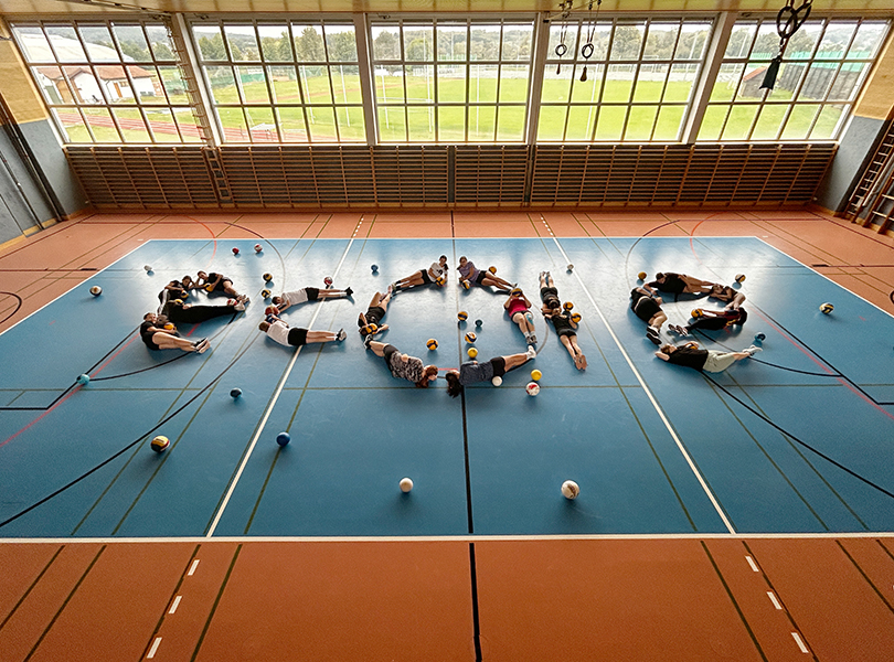 Schüler liegen am Boden vom Turnsaal und bilden das Wort ecole